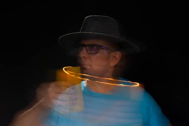 Mavi tişörtlü, sakallı, kel, şapkalı, yanan mumla ışık saçan adamın sanatsal portresi. Hareket bulanıklığı. Siyah arkaplan.