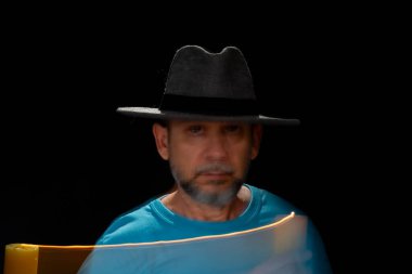 Mavi tişörtlü, sakallı, kel, şapkalı, yanan mumla ışık saçan adamın sanatsal portresi. Hareket bulanıklığı. Siyah arkaplan.