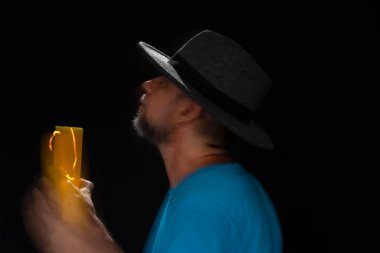 Mavi tişörtlü, sakallı, kel, şapkalı, yanan mumla ışık saçan adamın sanatsal portresi. Hareket bulanıklığı. Siyah arkaplan.