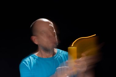 Mavi gömlekli kel adamın sanatsal portresi yanan sarı mumla hareket bulanıklığı yaratıyor. Hafif patika. Siyah arkaplan.