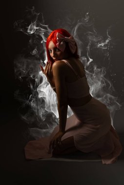 Pembe elbiseli genç, kızıl saçlı kadın yere çömelmiş, karanlık arka planda fotoğraf için poz veriyor. Stüdyo resmi