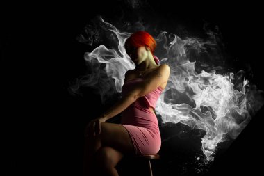 Güzel, genç, kızıl saçlı bir kadının portresi, oturan, pembe elbiseli, koyu arkaplanda fotoğraf için poz veren. Stüdyo resmi.