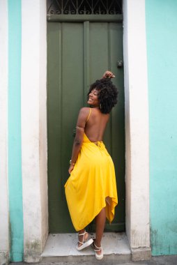 Afro saçlı, sarı elbiseli, yeşil kapıya dayanmış fotoğraf için poz veren güzel siyahi kadın portresi. Yolculukta mutlu insan.