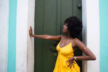 Afro saçlı, sarı elbiseli, yeşil kapıya dayanmış fotoğraf için poz veren güzel siyahi kadın portresi. Yolculukta mutlu insan.