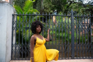 Eski demir bir çite dayanan sarı bir elbise giyen afro saçlı güzel siyah bir kadının portresi. Yolculuktaki mutlu insan..