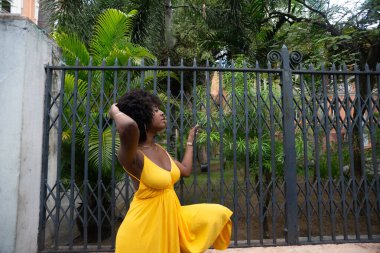 Eski demir bir çite dayanan sarı bir elbise giyen afro saçlı güzel siyah bir kadının portresi. Yolculuktaki mutlu insan..