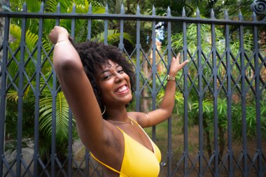 Eski demir bir çite dayanan sarı bir elbise giyen afro saçlı güzel siyah bir kadının portresi. Yolculuktaki mutlu insan..