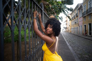 Eski demir bir çite dayanan sarı bir elbise giyen afro saçlı güzel siyah bir kadının portresi. Yolculuktaki mutlu insan..