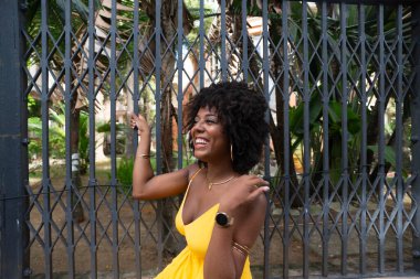 Afro saçlı, sarı elbiseli, mutlu bir kadın eski demir bir çite yaslanmış kameraya poz veriyor. Yolculuktaki mutlu insan..