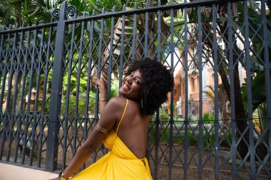 Afro saçlı, sarı elbiseli, mutlu bir kadın eski demir bir çite yaslanmış kameraya poz veriyor. Yolculuktaki mutlu insan..