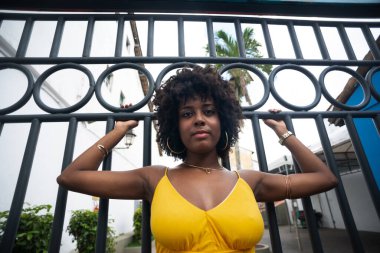 Demir bir çitin yanında sarı elbiseli, afro saçlı bir kadın. Kendinden emin ve mutlu biri.