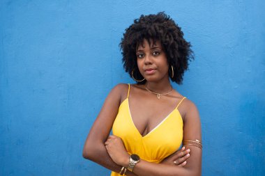 Sarı elbiseli, afro saçlı, mavi renkli bir duvarda izole edilmiş güzel bir kadın. Kendinden emin ve mutlu biri.