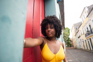 Sarı elbiseli, afro saçlı güzel bir kadın kırmızı kapının yanında fotoğraf çektirmek için poz veriyor. Kendinden emin ve mutlu biri. Brezilya