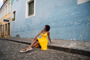 Sarı elbiseli ve afro saçlı güzel bir kadının portresi mavi duvara karşı eski caddenin kaldırımında oturuyor. Kendinden emin ve mutlu biri. Brezilya