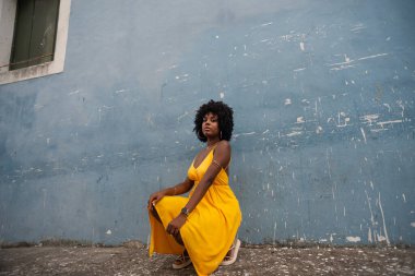 Sarı elbiseli güzel bir kadının portresi ve mavi bir evin duvarına çömelmiş afro saç modeli. Kendinden emin ve mutlu biri. Brezilya