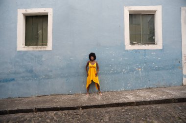 Sarı elbiseli ve afro saçlı güzel bir kadın pencereler arasındaki evin duvarına yaslanmış bir fotoğraf için poz veriyor. Kendinden emin ve mutlu biri. Brezilya