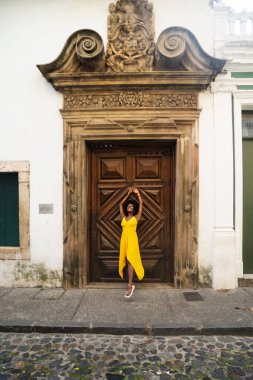 Sarı elbiseli, afro saçlı, yüzyıllık ahşap kapıya dayanmış güzel bir genç kadın. Kendine güvenen biri. Brezilya