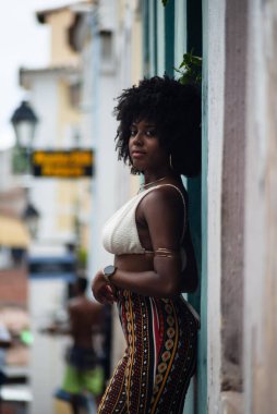 Arkası duvara dönük, fotoğraf için poz veren güzel siyahi bir kadının yarım boy fotoğrafı. Gülümseyen ve güçlü bir insan. Brezilya