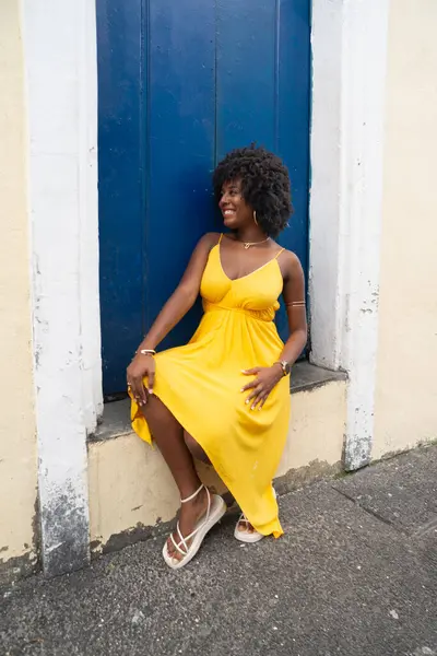 Afro saçlı, sarı elbiseli, eski bir evin mavi kapısında oturan güzel bir genç kadın. Kendine güvenen biri. Brezilya