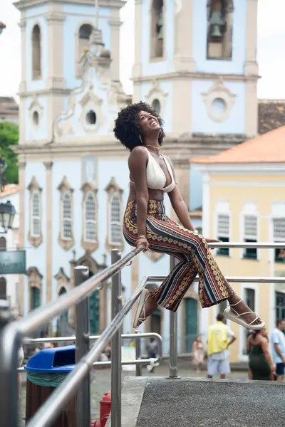 Siyah saçlı genç bir kadın paslanmaz çelikten bir korkulukta oturuyor ve arka planda evler var. Mutlu ve güçlü biri. Brezilya