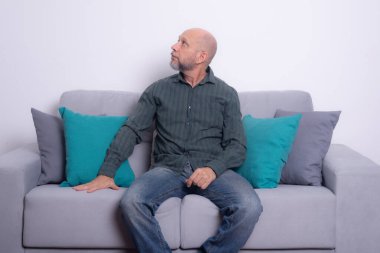 Uzun kollu gömlek ve kot pantolon giyen sakallı kel adam gri bir koltukta oturuyor, rahatlamış, yukarı bakıyor. Gri bir arkaplanda.