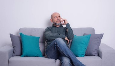 Uzun kollu gömlek ve kot pantolon giyen sakallı kel bir adam gri bir kanepeye oturmuş poz veriyor ve yukarı bakıyor. Gri bir arkaplanda.