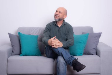 Uzun kollu gömlek ve kot pantolon giyen sakallı kel bir adam gri bir kanepeye oturmuş poz veriyor ve yukarı bakıyor. Gri bir arkaplanda.