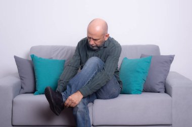 Uzun kollu gömlekli ve kot pantolonlu sakallı kel bir adam kanepeye oturmuş, ayakkabısını giyiyor. Gri bir arkaplanda.