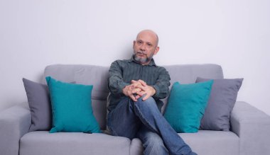 Uzun kollu gömlek ve kot pantolon giymiş rahat, kel, sakallı bir adam gri bir kanepeye oturmuş fotoğrafa bakıyor. Gri bir arkaplanda.