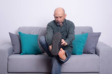 Uzun kollu gömlekli ve kot pantolonlu sakallı kel bir adam kanepeye oturmuş, ayakkabısını giyiyor. Gri bir arkaplanda.