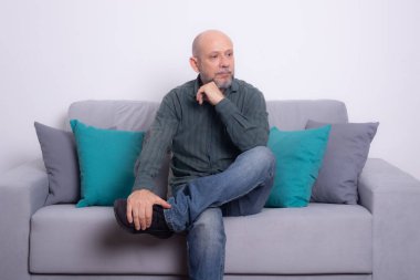 Uzun kollu gömlek ve kot pantolon giyen sakallı kel adam kanepede oturuyor. Cidden aşağıya bakıyorum. Gri bir arkaplanda.