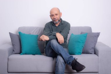 Uzun kollu gömlek ve kot pantolon giyen kel, sakallı bir adam bir kanepeye oturur ve bacaklarını çaprazlar, yan tarafa bakar. Gri bir arkaplanda.