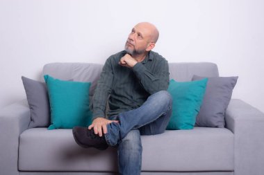 Uzun kollu gömlek ve kot pantolon giyen sakallı kel bir adam gri bir kanepeye oturmuş poz veriyor ve yukarı bakıyor. Gri bir arkaplanda.