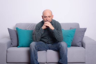 Uzun kollu gömlek ve kot pantolon giyen sakallı kel adam kanepede oturuyor. Cidden aşağıya bakıyorum. Gri bir arkaplanda.