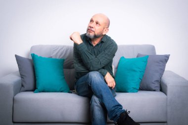 Uzun kollu gömlek ve kot pantolon giyen sakallı kel bir adam gri bir kanepeye oturmuş poz veriyor ve yukarı bakıyor. Gri bir arkaplanda.