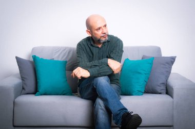 Uzun kollu gömlek ve kot pantolon giyen kel, sakallı bir adam bir kanepeye oturur ve bacaklarını çaprazlar, yan tarafa bakar. Gri bir arkaplanda.