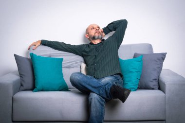 Uzun kollu gömlek ve kot pantolon giyen sakallı kel bir adam gri bir kanepeye oturmuş poz veriyor ve yukarı bakıyor. Gri bir arkaplanda.