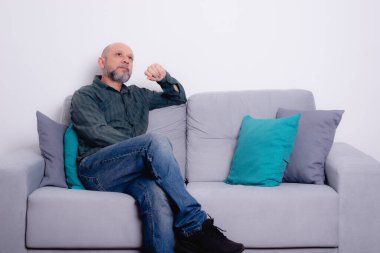 Uzun kollu gömlek ve kot pantolon giyen sakallı kel bir adam gri bir kanepeye oturmuş poz veriyor ve yukarı bakıyor. Gri bir arkaplanda.