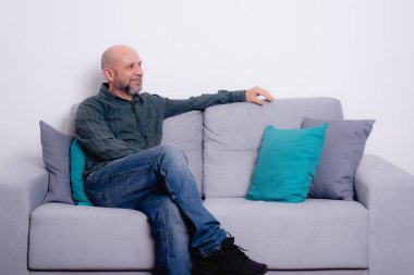 Uzun kollu gömlek ve kot pantolon giyen kel, sakallı bir adam bir kanepeye oturur ve bacaklarını çaprazlar, yan tarafa bakar. Gri bir arkaplanda.