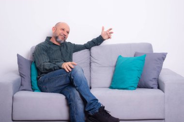 Uzun kollu gömlek ve kot pantolon giyen sakallı kel adam gri bir koltukta oturuyor, rahatlamış, yukarı bakıyor. Gri bir arkaplanda.