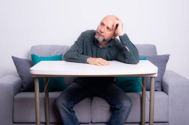 Kel, iyi giyimli, sakallı bir adam, kollarını masaya uzatmış, düşünceli ve sakin bir şekilde oturuyor. Beyaz arkaplana karşı.