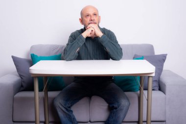 Kel, iyi giyimli, sakallı bir adam düşünceli bir şekilde koltukta oturuyor ve kolları masanın üzerinde dinleniyor. Beyaz arkaplana karşı.