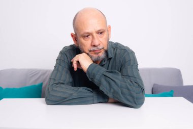 Ciddi, resmi tişörtlü, sakallı, kollarını masaya uzatmış, düşünceli ve sakin bir adam. Beyaz arkaplana karşı.