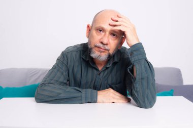 Kel, iyi giyimli, sakallı bir adam, kollarını masaya uzatmış, düşünceli ve sakin bir şekilde oturuyor. Beyaz arkaplana karşı.