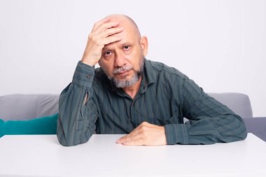 Kel, iyi giyimli, sakallı bir adam, kollarını masaya uzatmış, düşünceli ve sakin bir şekilde oturuyor. Beyaz arkaplana karşı.