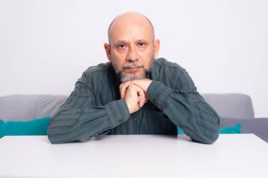 Kel, iyi giyimli, sakallı bir adam düşünceli bir şekilde koltukta oturuyor ve kolları masanın üzerinde dinleniyor. Beyaz arkaplana karşı.