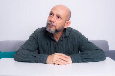 Kel, iyi giyimli, sakallı bir adam koltukta oturuyor, kafası masaya eğik, derin düşüncelere dalmış. Beyaz arkaplana karşı.