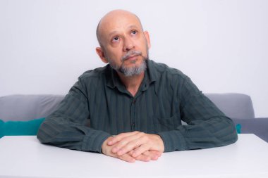Ciddi, resmi tişörtlü, sakallı, kollarını masaya uzatmış, düşünceli ve sakin bir adam. Beyaz arkaplana karşı.