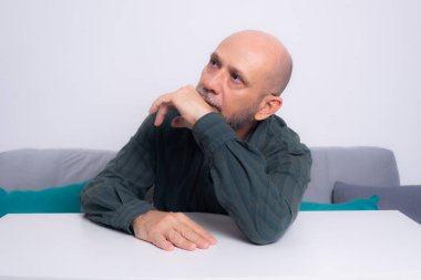 Ciddi, resmi tişörtlü, sakallı, kollarını masaya uzatmış, düşünceli ve sakin bir adam. Beyaz arkaplana karşı.