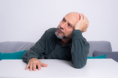 Kel, iyi giyimli, sakallı bir adam, kollarını masaya uzatmış, düşünceli ve sakin bir şekilde oturuyor. Beyaz arkaplana karşı.